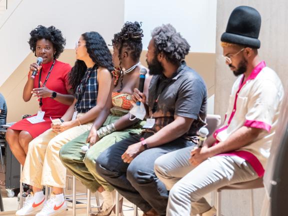 Five panelists with microphones sitting on tall chairs on a stage