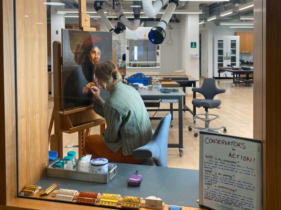 An MFA conservator holding a swab against a painting during treatment as seen through the glass windows of the Conservation Center