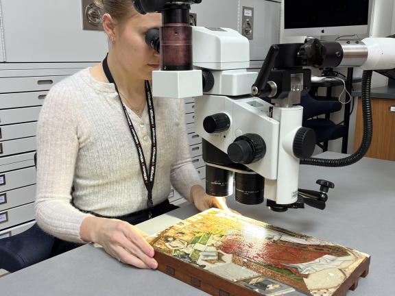 A light from a microscope shines on a small panel painting while an MFA conservators looks through the microscope