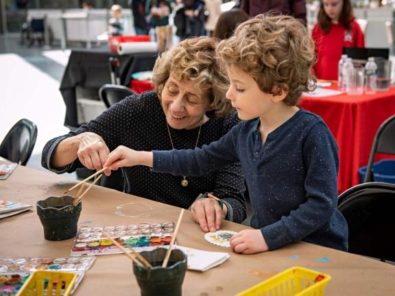A young child and an older woman sit side by side at a table covered with kraft paper and work on an art-making activity 